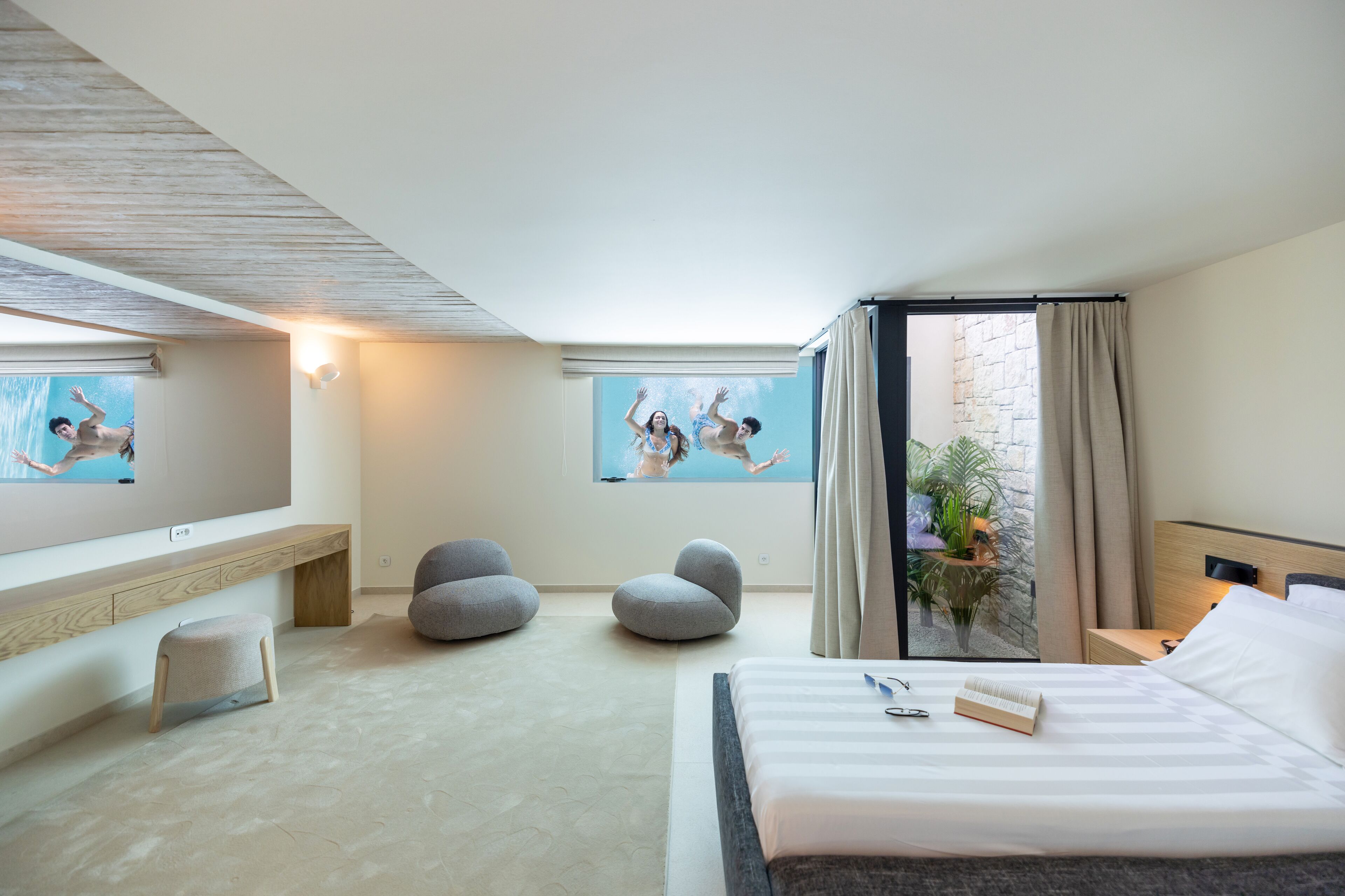 A modern hotel room with a large bed, a flat-screen TV, a balcony with a view, and a small seating area with bean bags.