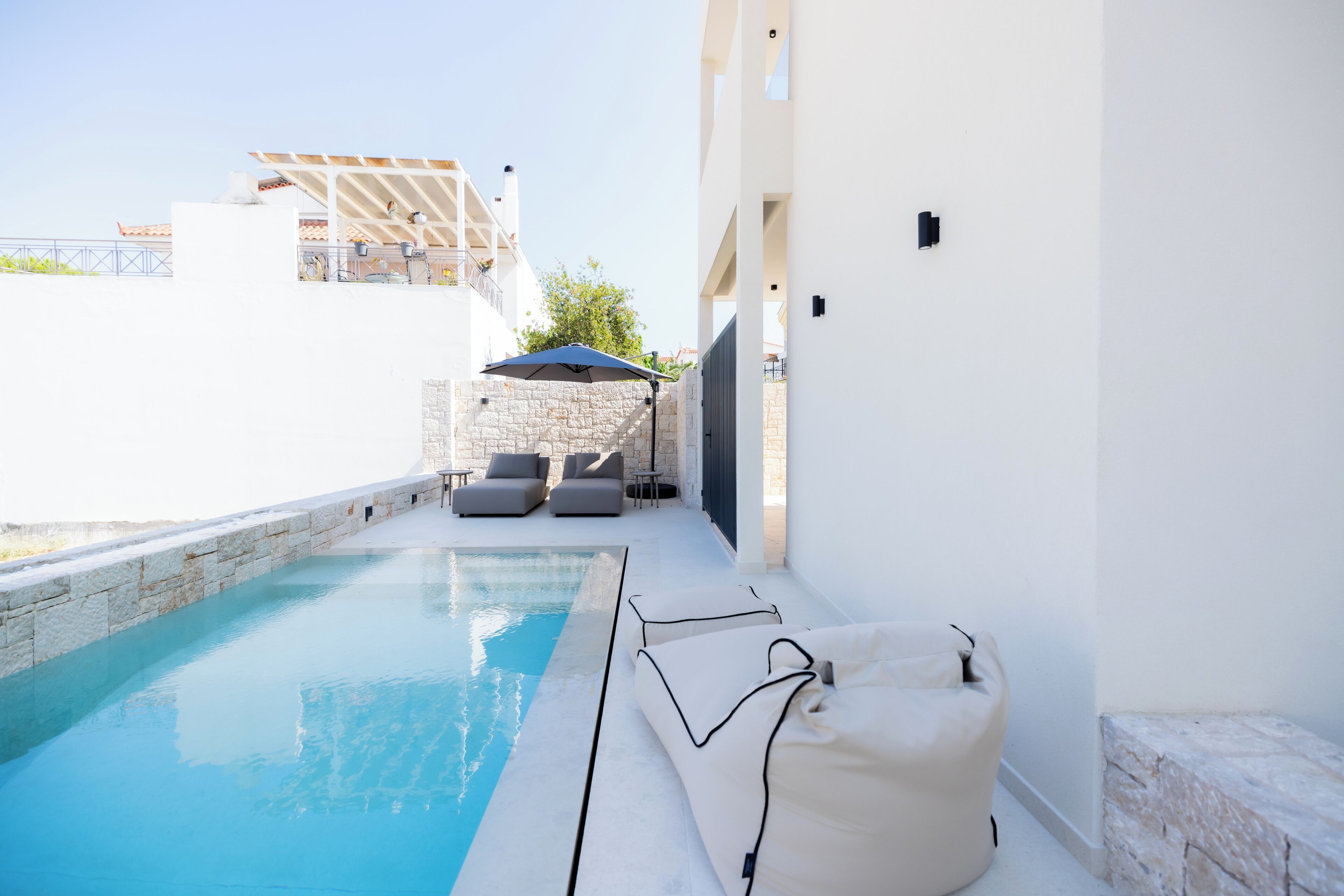 A pool area with a stone wall, a white building, and a covered seating area with cushions.