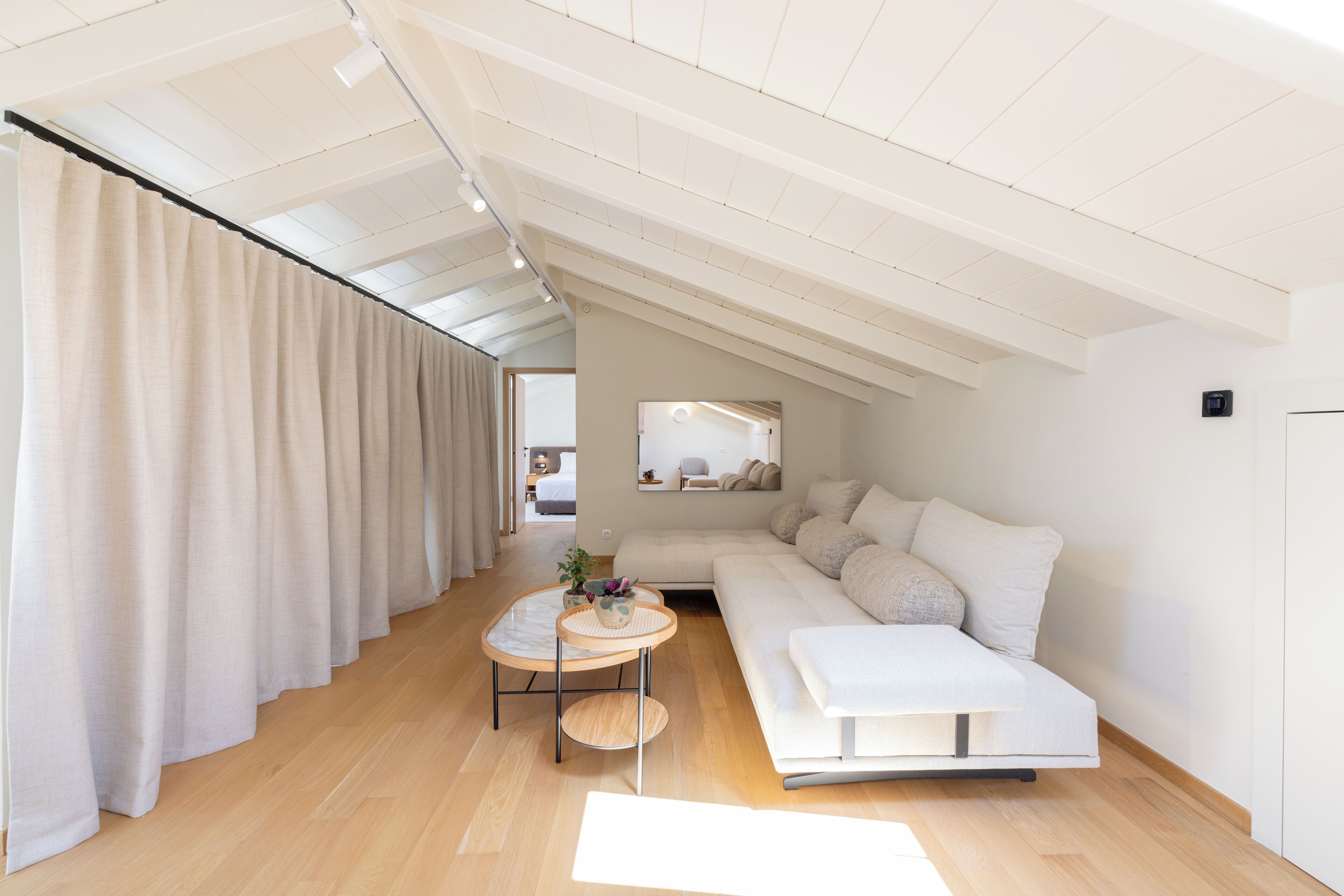 A modern loft bedroom with a high ceiling, a large window, a white sofa, a small round table, and a bed in the background.