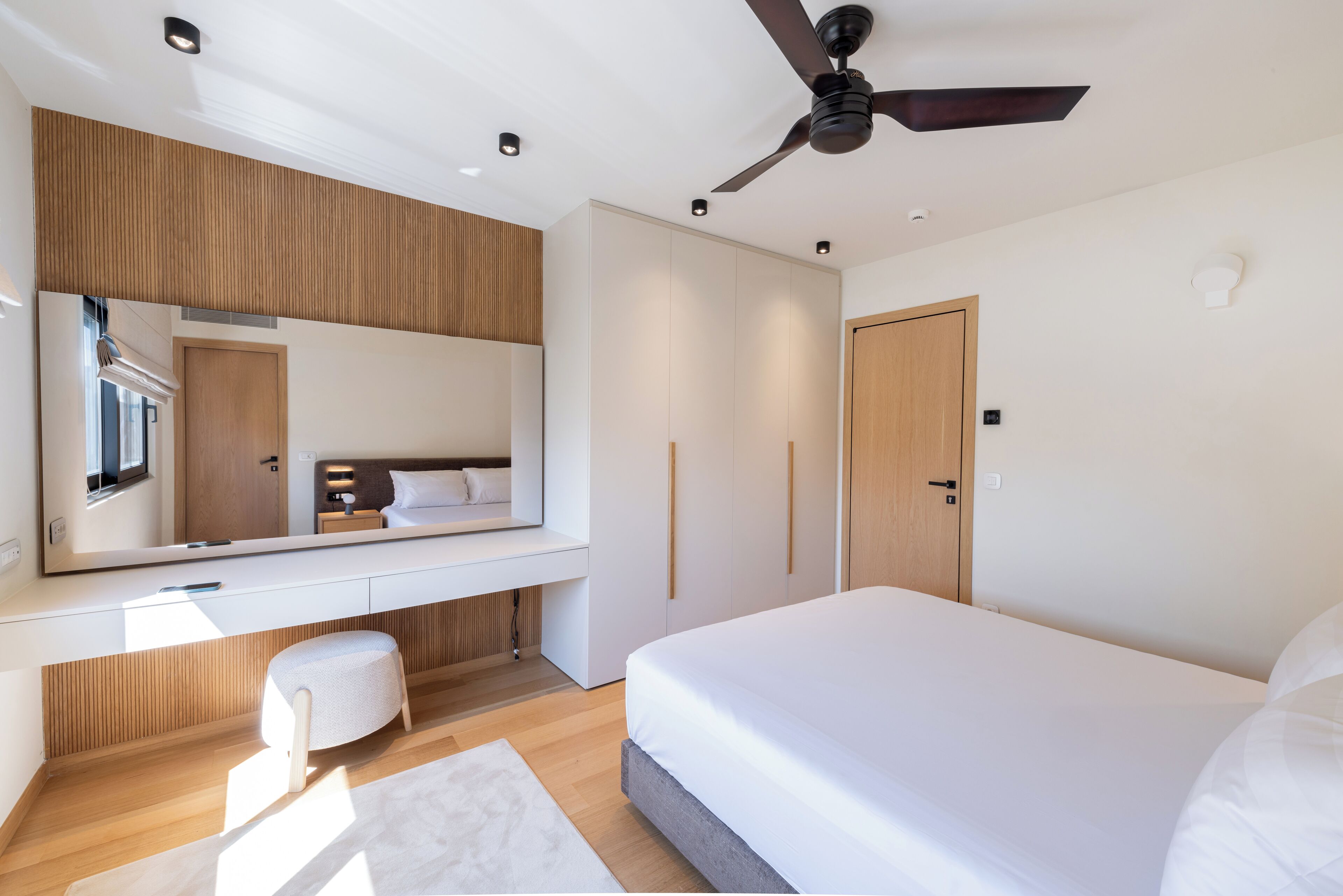 A modern bedroom with a large bed, a vanity with a mirror, a ceiling fan, and built-in wardrobes.