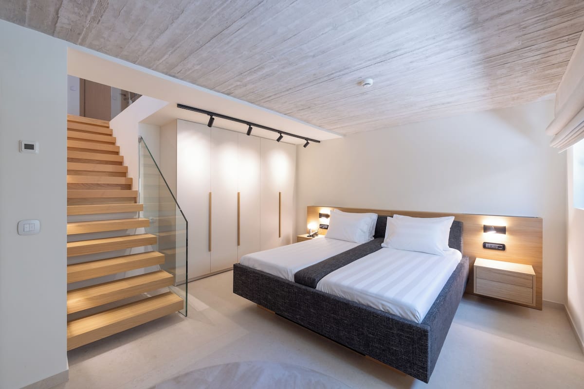 A modern bedroom with a large bed, a wooden staircase, and a glass railing.