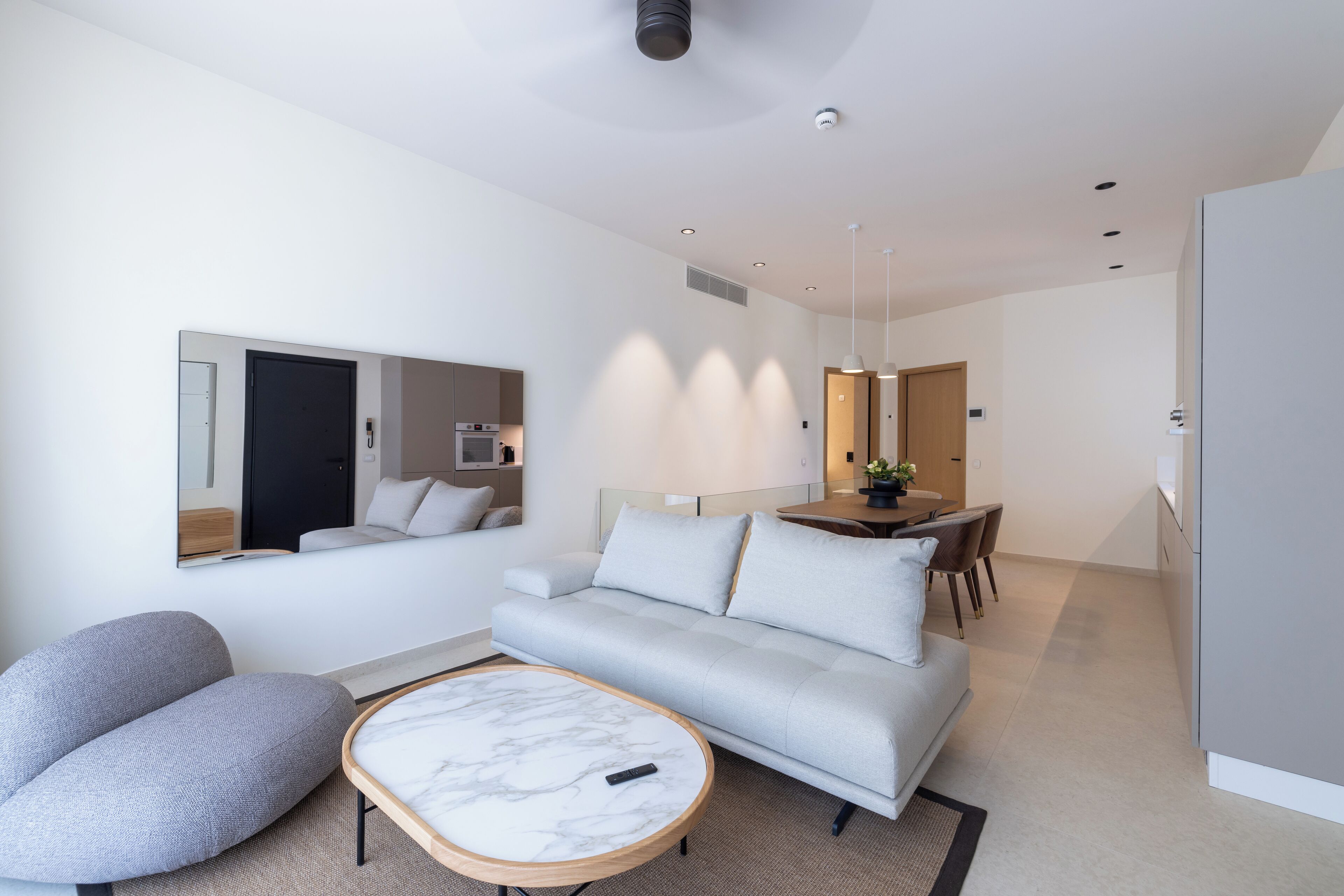 A modern living room with a sofa, armchair, and a marble coffee table. There is a dining area with a table and chairs in the background.