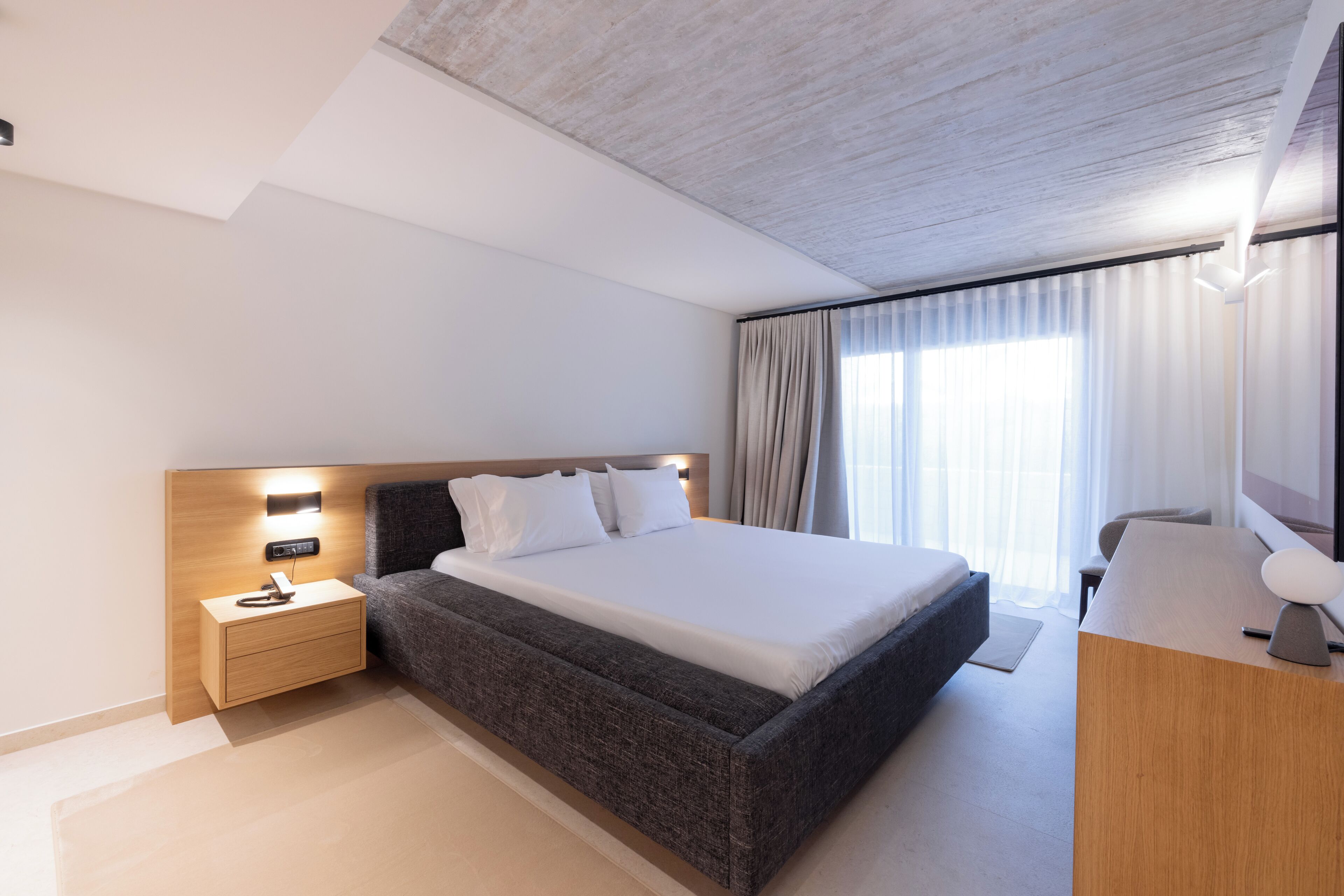 Una habitación de hotel moderna con una cama grande, una mesita de noche con una lámpara, una mesita auxiliar de madera y un escritorio con una silla.