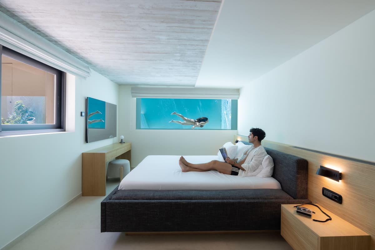 A modern bedroom with a large bed, a bedside table with a lamp, a window with a view of the sky, and a wall-mounted artwork.