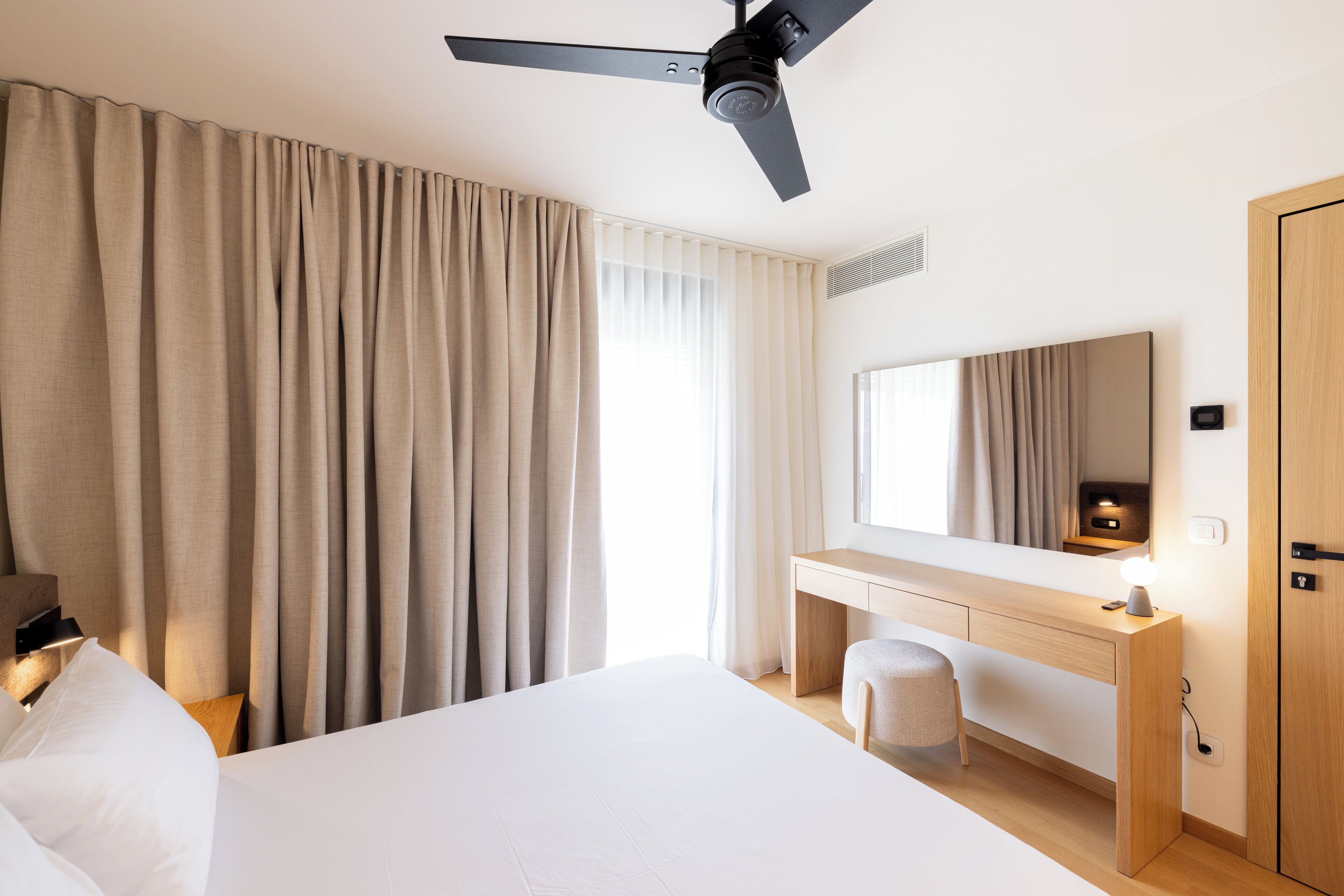 Habitación de hotel con cama, escritorio, silla, ventilador de techo y un espejo grande.