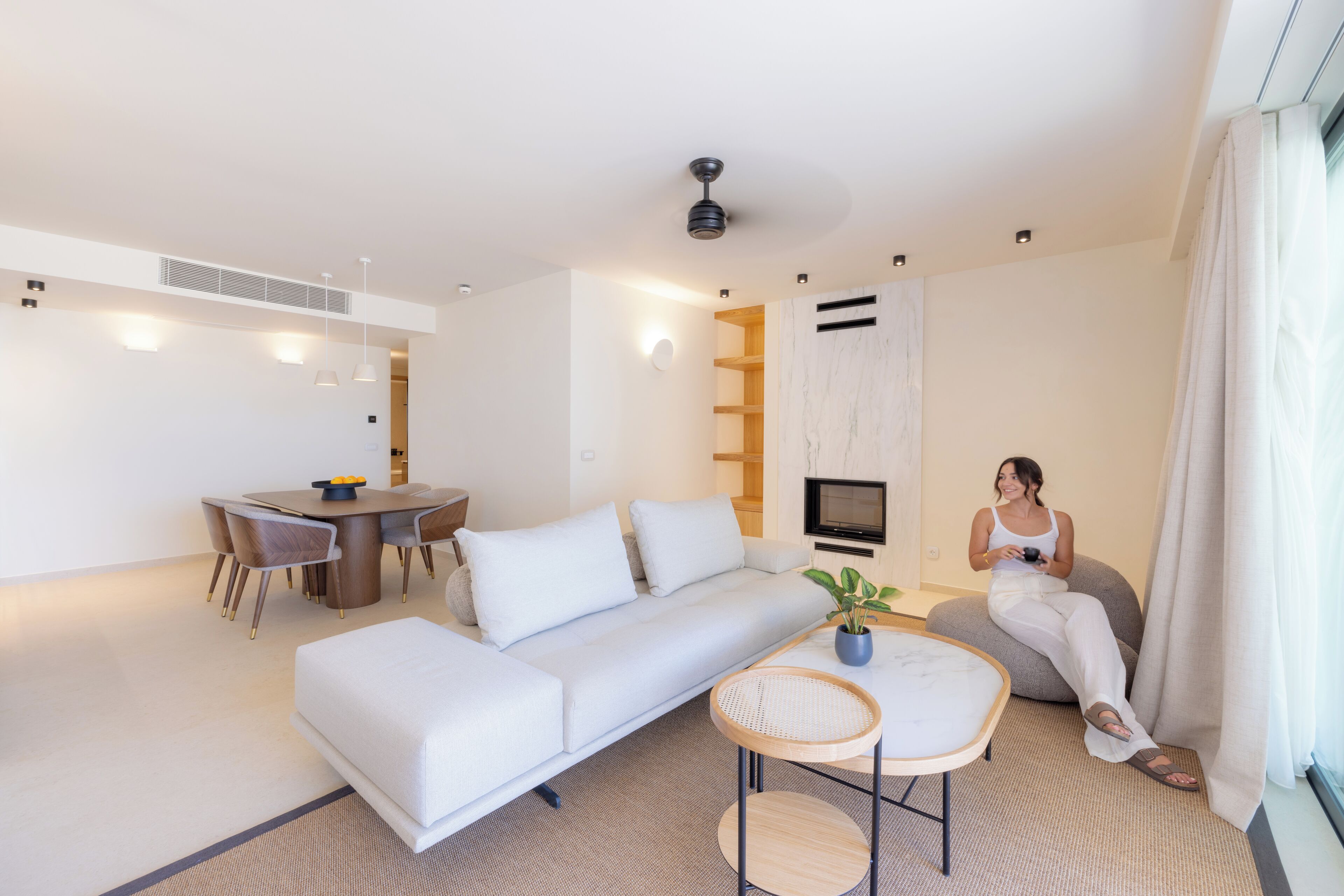 A modern living room with a white sofa, dining table, and chairs. There is a TV mounted on the wall, a ceiling fan, and a potted plant.