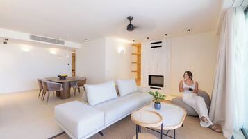 A modern living room with a white sofa, dining table, and chairs. There is a TV mounted on the wall, a ceiling fan, and a potted plant.