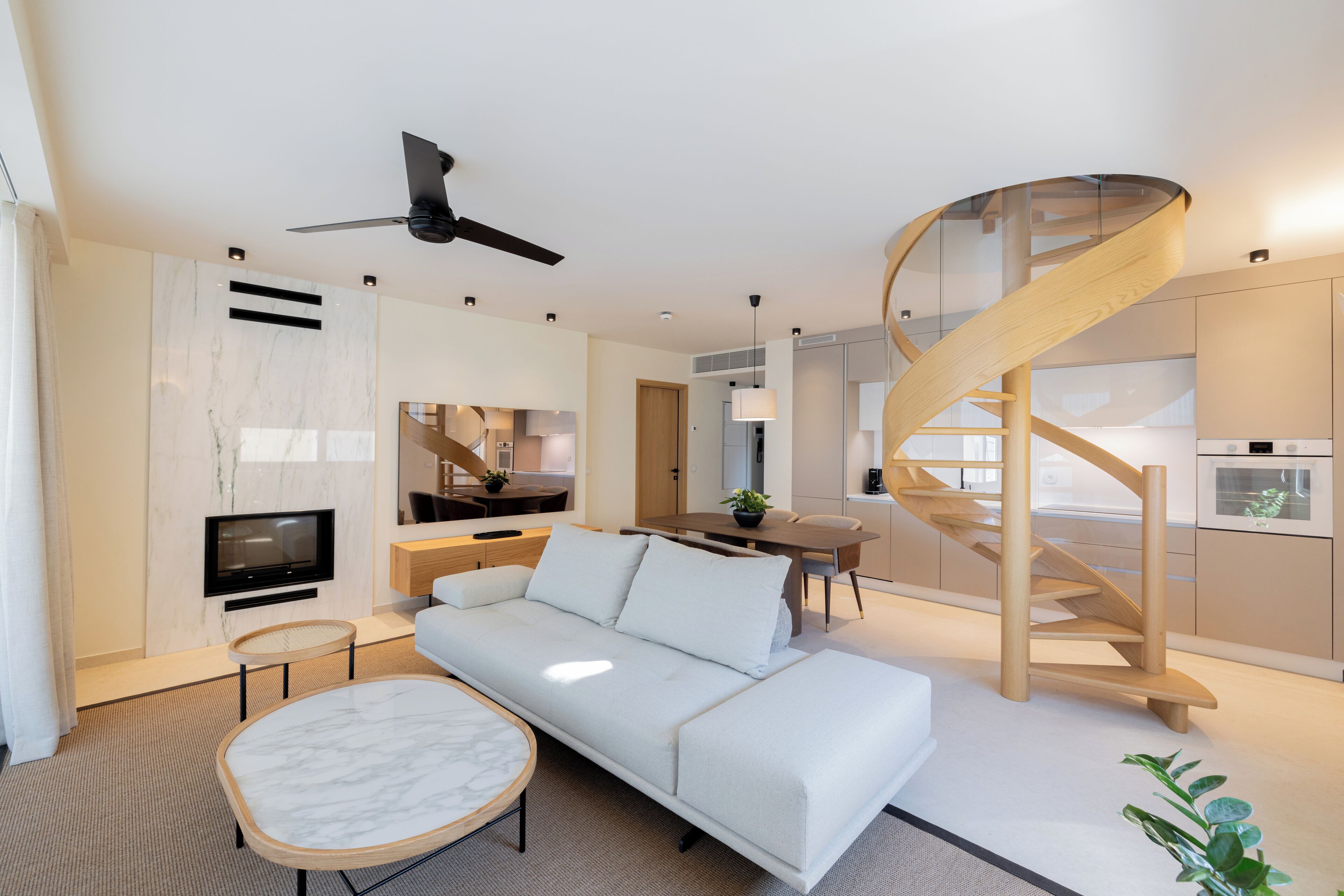 A modern living room with a white sofa, a round coffee table, a TV mounted on the wall, a ceiling fan, and a spiral staircase leading to an upper level.