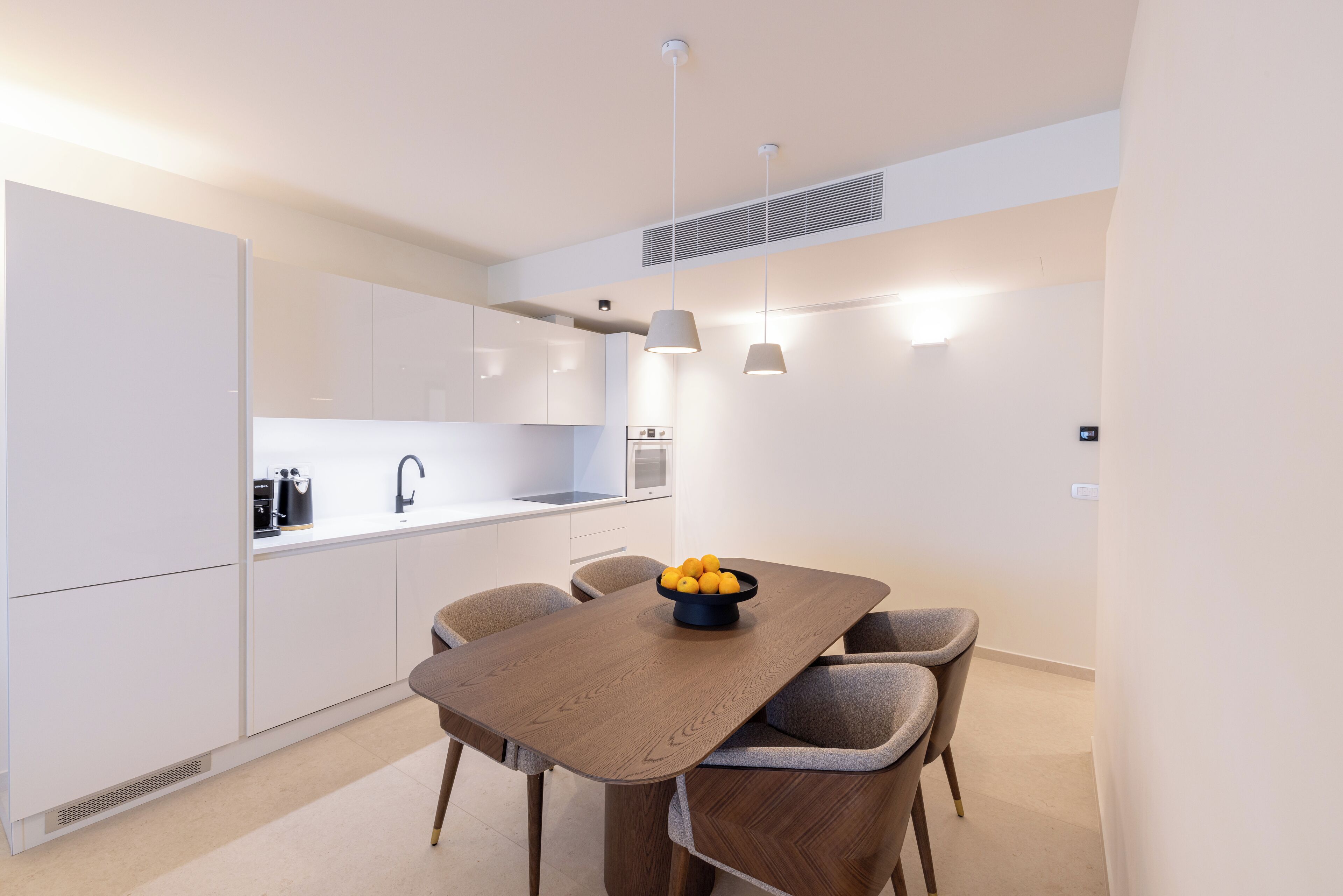 A modern kitchen with white cabinets, a dark wooden dining table, and a bowl of fruit.