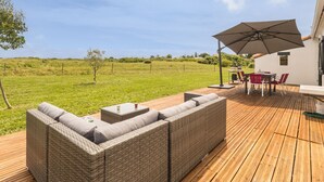 A covered outdoor seating area with a wicker sofa, a table, and chairs, overlooking a grassy field.