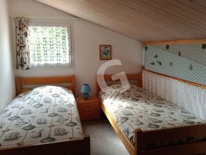Un dormitorio con dos camas individuales, una ventana con cortinas, una mesita de noche con lámpara y una estantería en la pared.