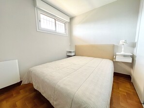 Een slaapkamer met een enkel bed, een nachtkastje, een bijzettafeltje met een lamp, een raam met jaloezieën en een radiator.