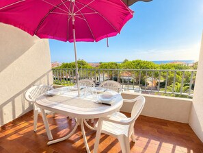 Een balkon met een tafel, stoelen, een roze parasol en uitzicht op zee en groen.