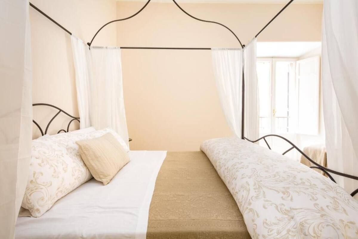 A double bed with a canopy, a bedside table, and a window with curtains.