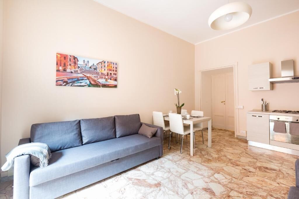 A modern living room with a grey sofa, a dining table with chairs, a kitchen area with a stove, and a painting of a street scene on the wall.