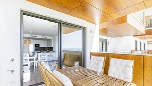 Ein moderner Essbereich mit Holztisch und weißen, gepolsterten Stühlen, im Hintergrund eine Küche und Blick auf das Meer durch große Glastüren.
