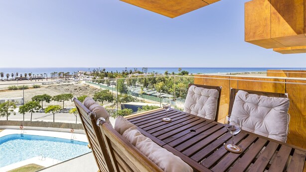 Eine Holzterrasse mit Pool, einem Tisch mit Kissen und Blick auf Strand und Meer.