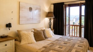A bedroom with a large bed, a nightstand, a lamp, a painting, and a view of the outdoors.