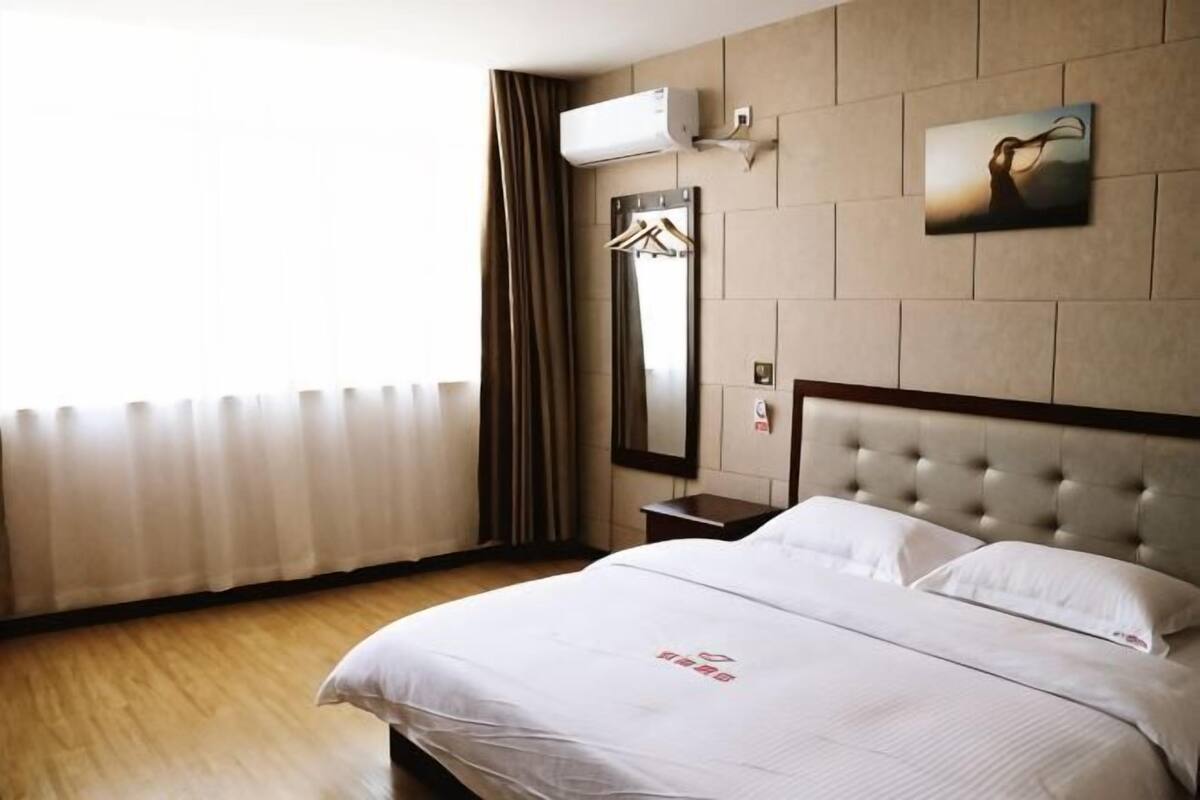 A hotel room with a bed, a mirror, a wall-mounted air conditioner, and a window with curtains.