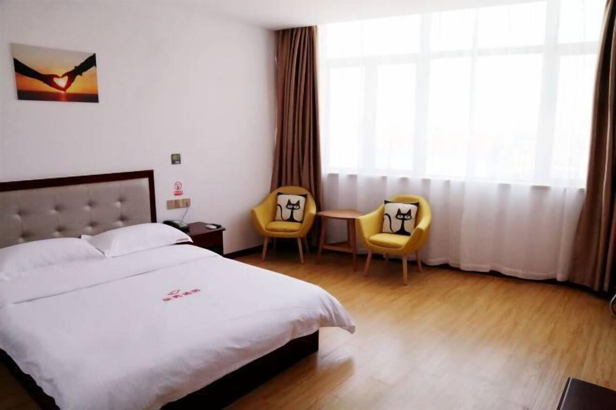 A hotel room with a bed, two yellow chairs, a small table, and a window with curtains.