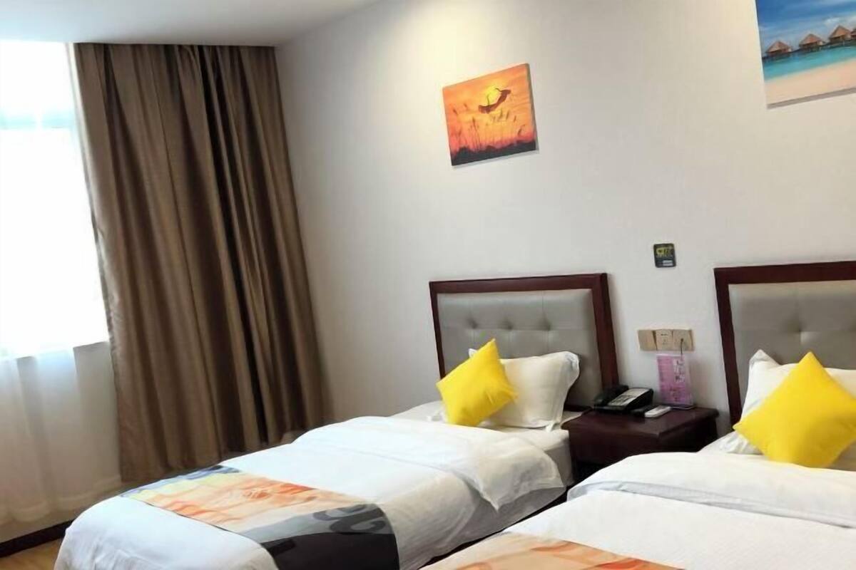 A hotel room with two beds, each with white bedding and yellow pillows, a nightstand with a phone, and a painting of a sunset on the wall.