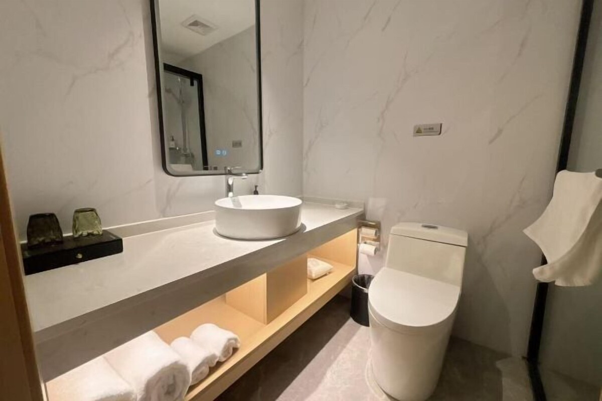 Ein modernes Badezimmer mit Waschbecken, Spiegel und Toilette. Handtücher liegen auf einem Regal, und ein Mülleimer steht bereit.