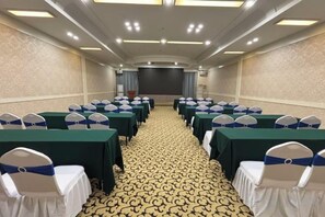 Un salón de conferencias con filas de mesas y sillas, piso alfombrado y una pantalla al frente.