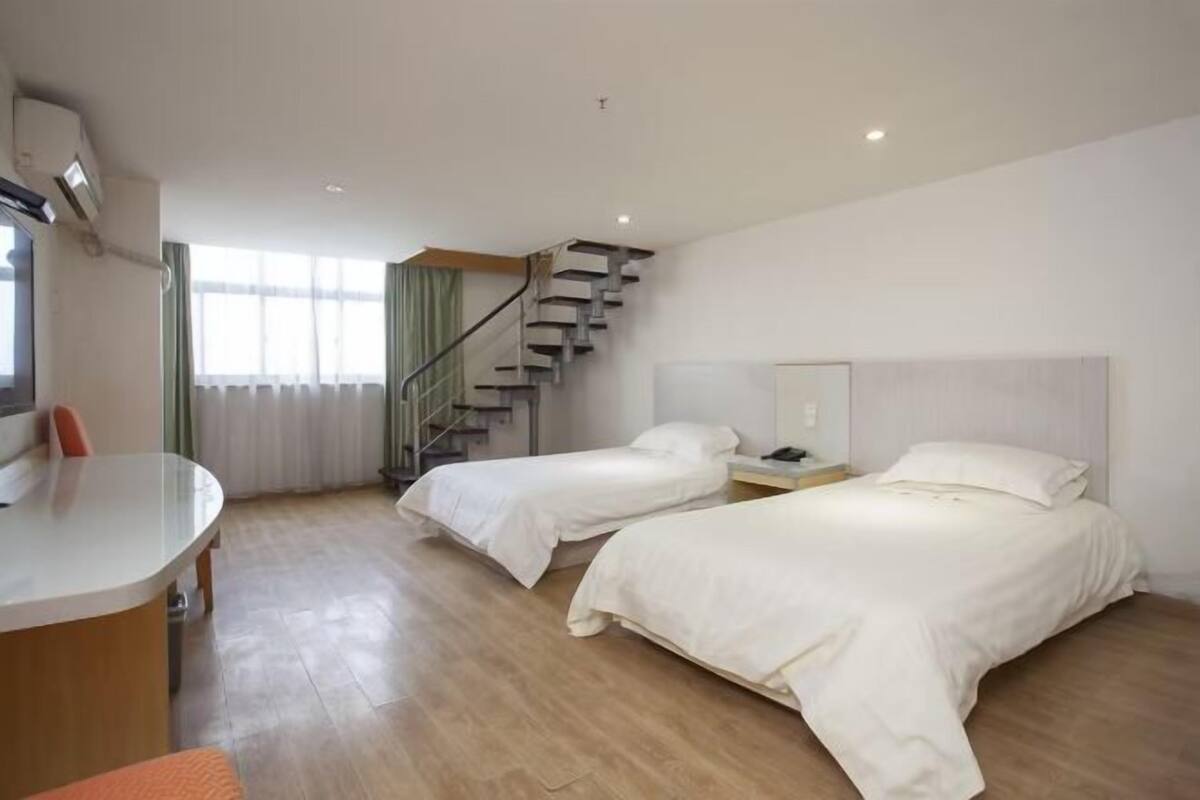 A hotel room with two beds, a desk, a chair, a staircase, and a window with curtains.