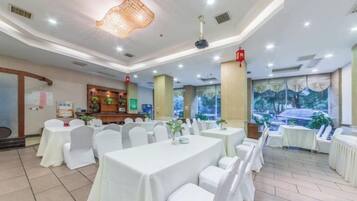 A banquet hall with round tables, white chairs, and a large window with a view of trees.