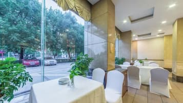 A hotel dining area with tables and chairs, large windows, and indoor plants.