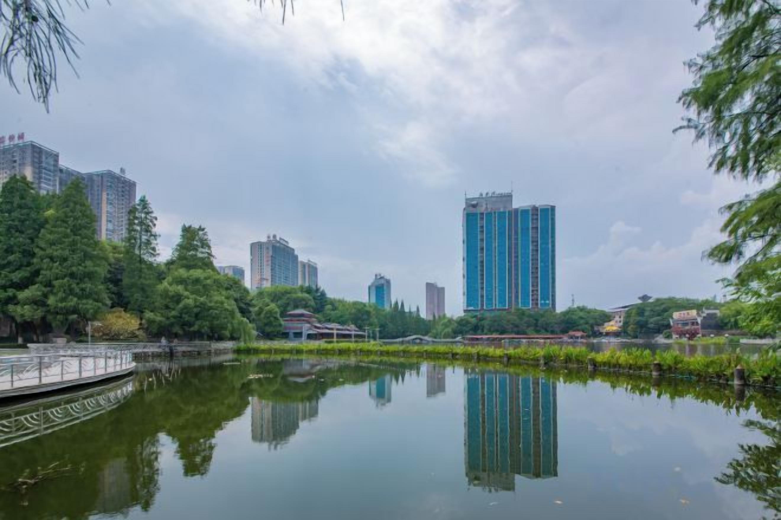 Um lago tranquilo refletindo edifícios, rodeado por árvores e uma ponte.