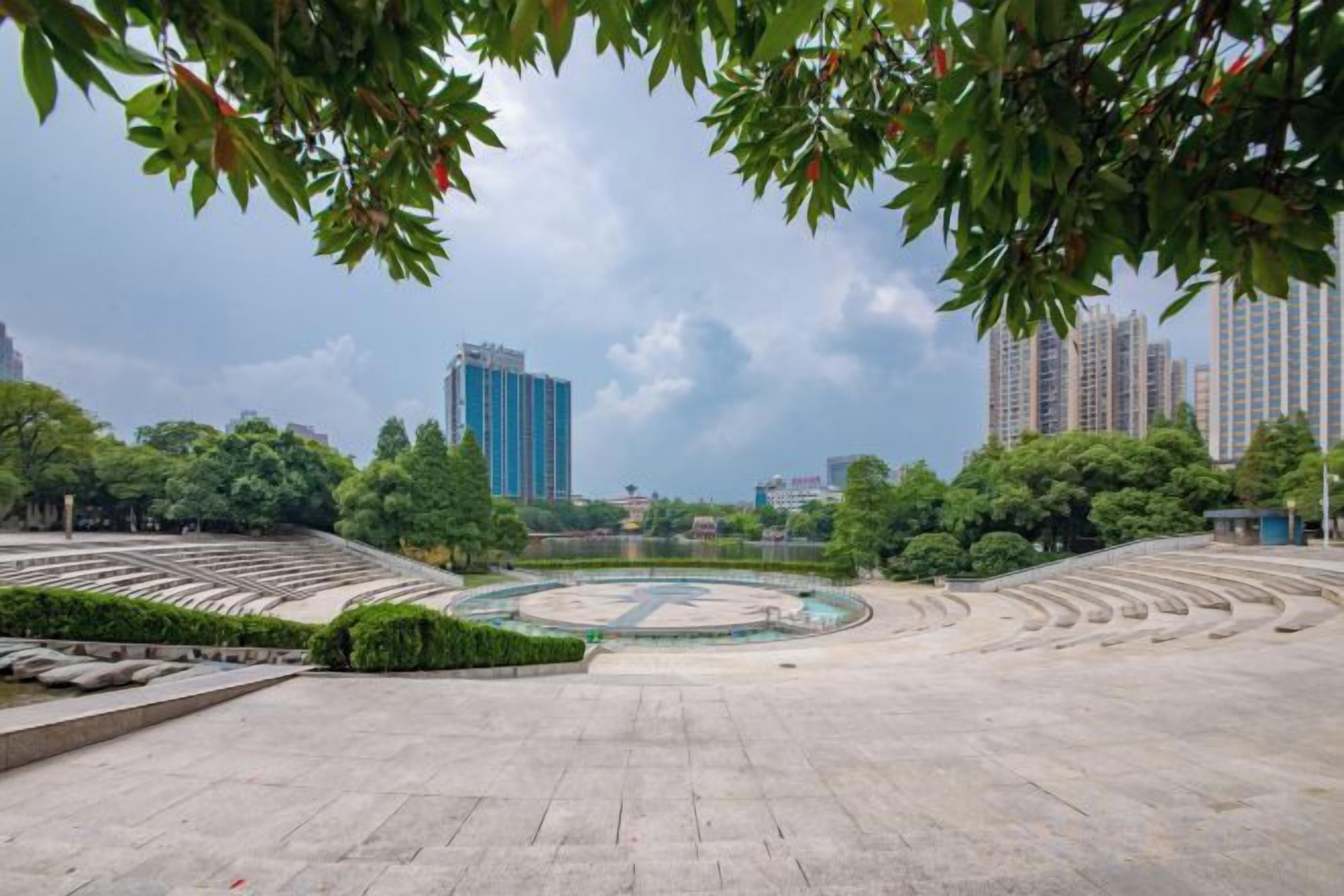 An open plaza with a circular fountain, surrounded by trees and buildings.
