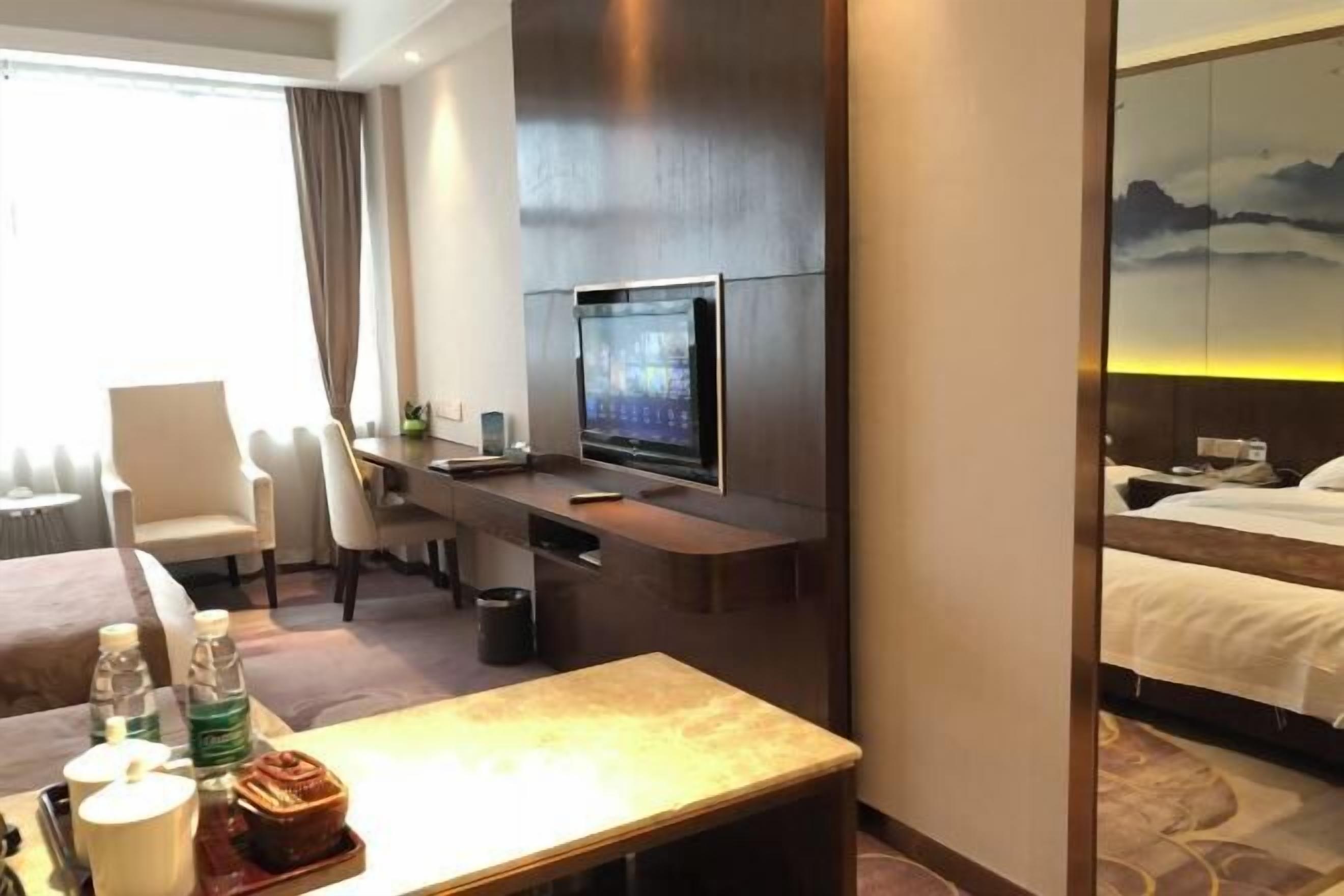 Ein Hotelzimmer mit einem Bett, einem Schreibtisch mit Fernseher, einem Sessel und einem Tisch mit Flasche und Tasse.