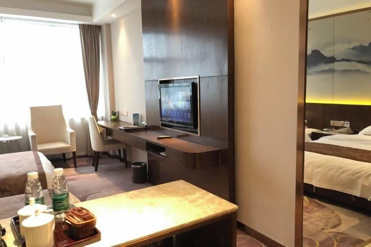A hotel room with a bed, a desk with a TV, a chair, and a table with a bottle and a cup.