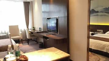 A hotel room with a bed, a desk with a TV, a chair, and a table with a bottle and a cup.
