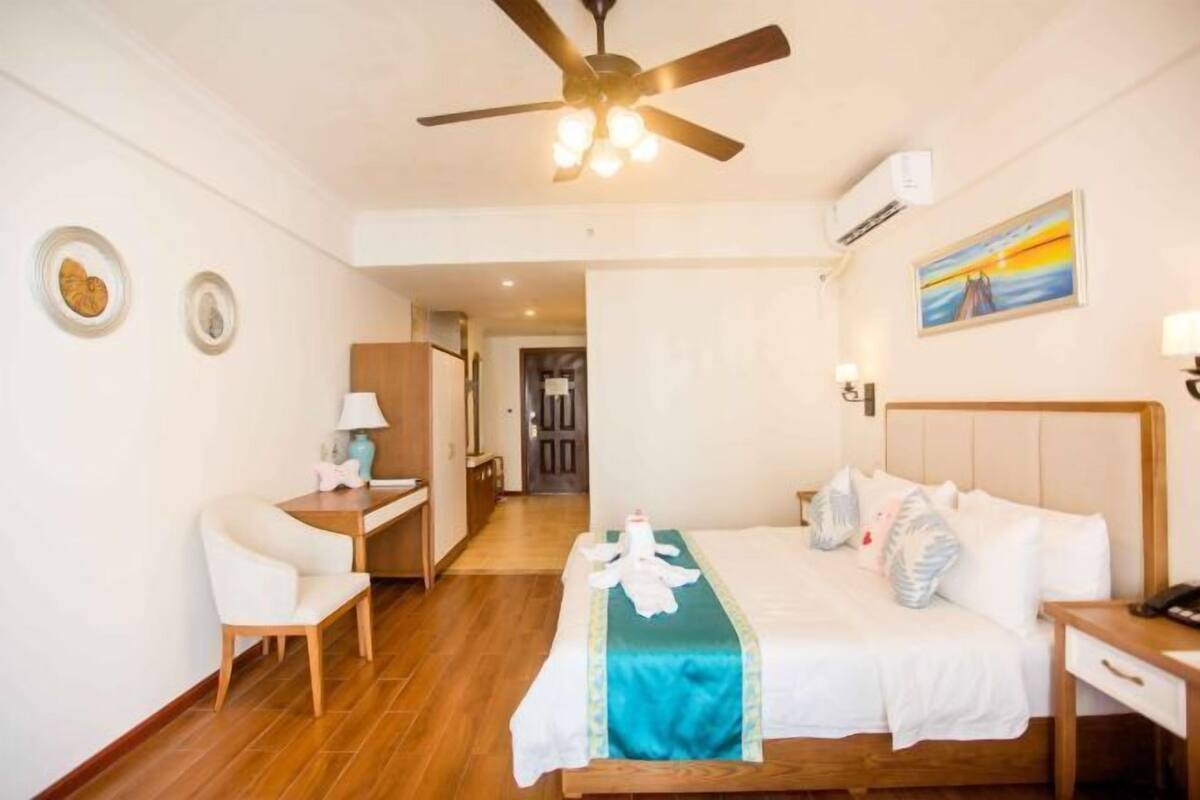 A hotel room with a bed, a desk, a chair, a ceiling fan, and a painting on the wall.