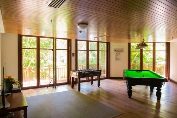 Una habitación con mesa de billar, mesa de futbolín y ventanas amplias con vistas a follaje.