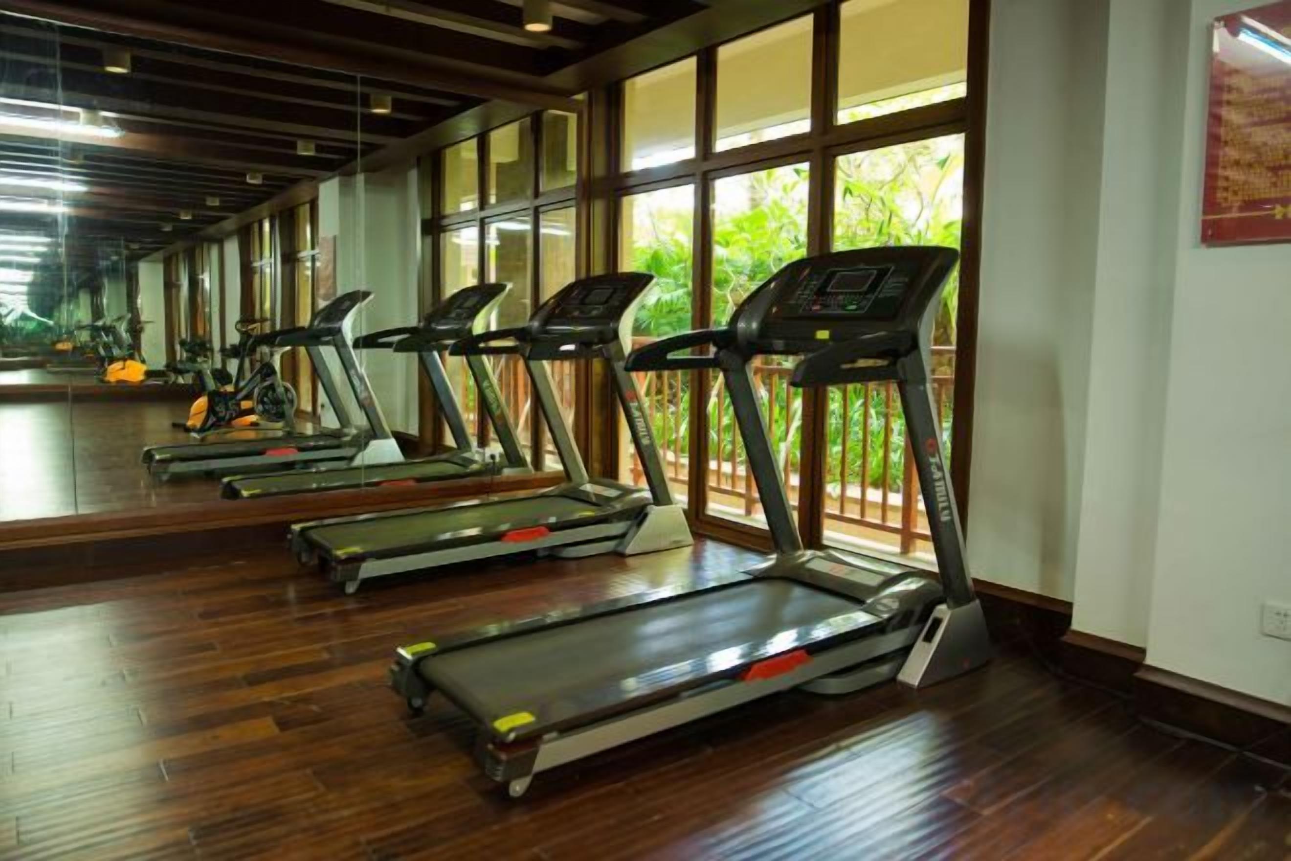 A row of treadmills in a gym with wooden flooring and large windows.