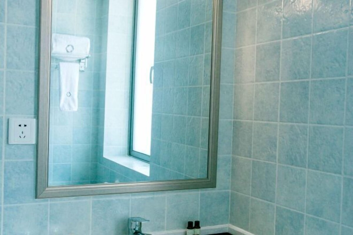 A bathroom with light blue tiles, a large mirror, a visible toilet paper holder, and a small shelf with toiletries.