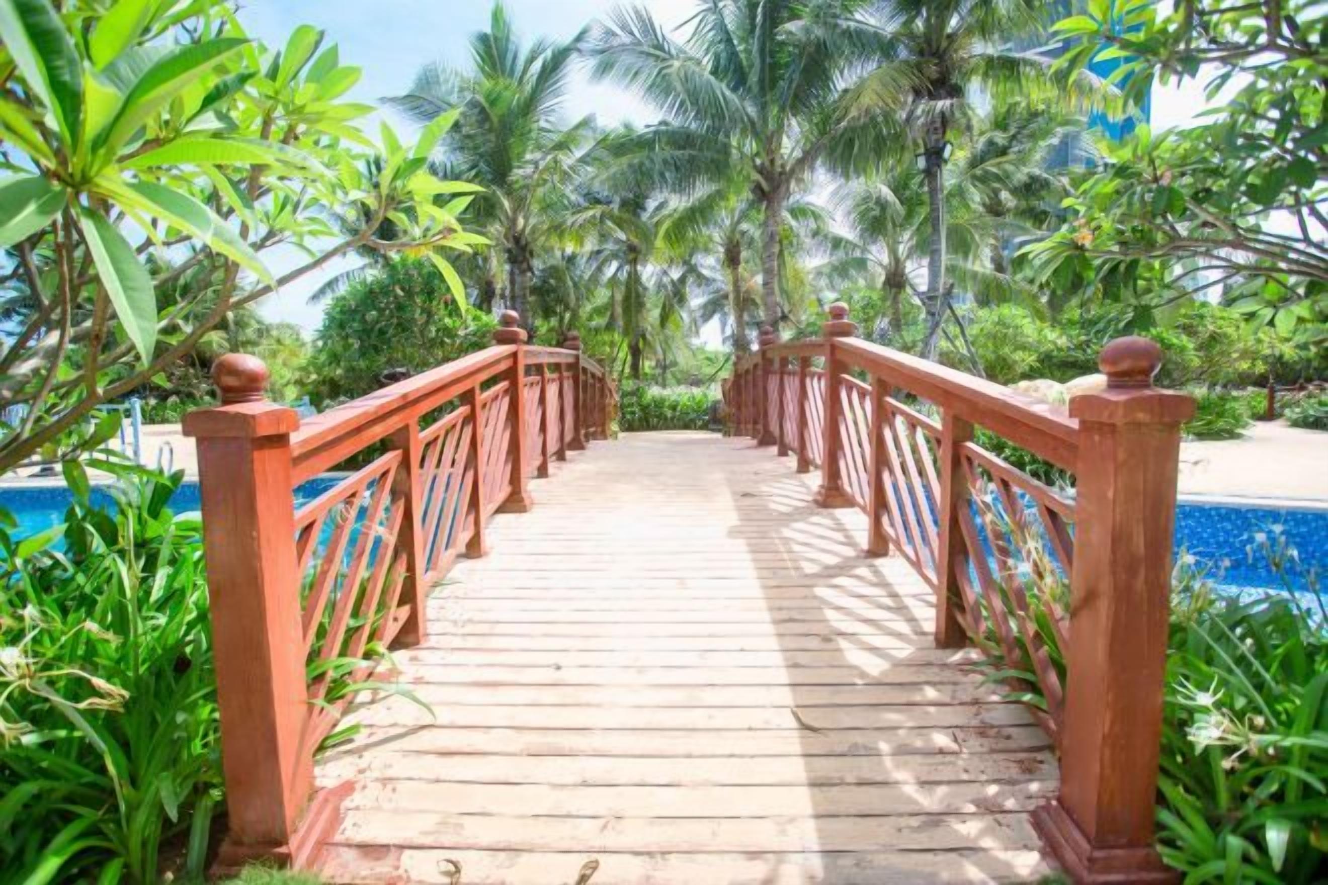 A wooden bridge leading to a pool area surrounded by lush greenery and palm trees.