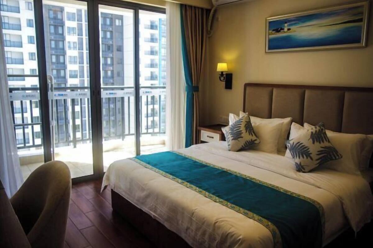 A hotel room with a large bed, a nightstand, a lamp, and a view of the city through a sliding door.
