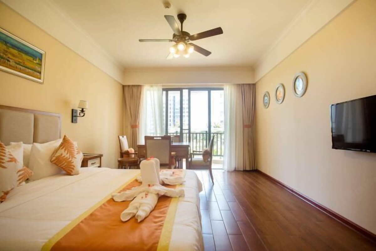 A hotel room with a bed, a ceiling fan, a TV, and a balcony with a table and chairs.