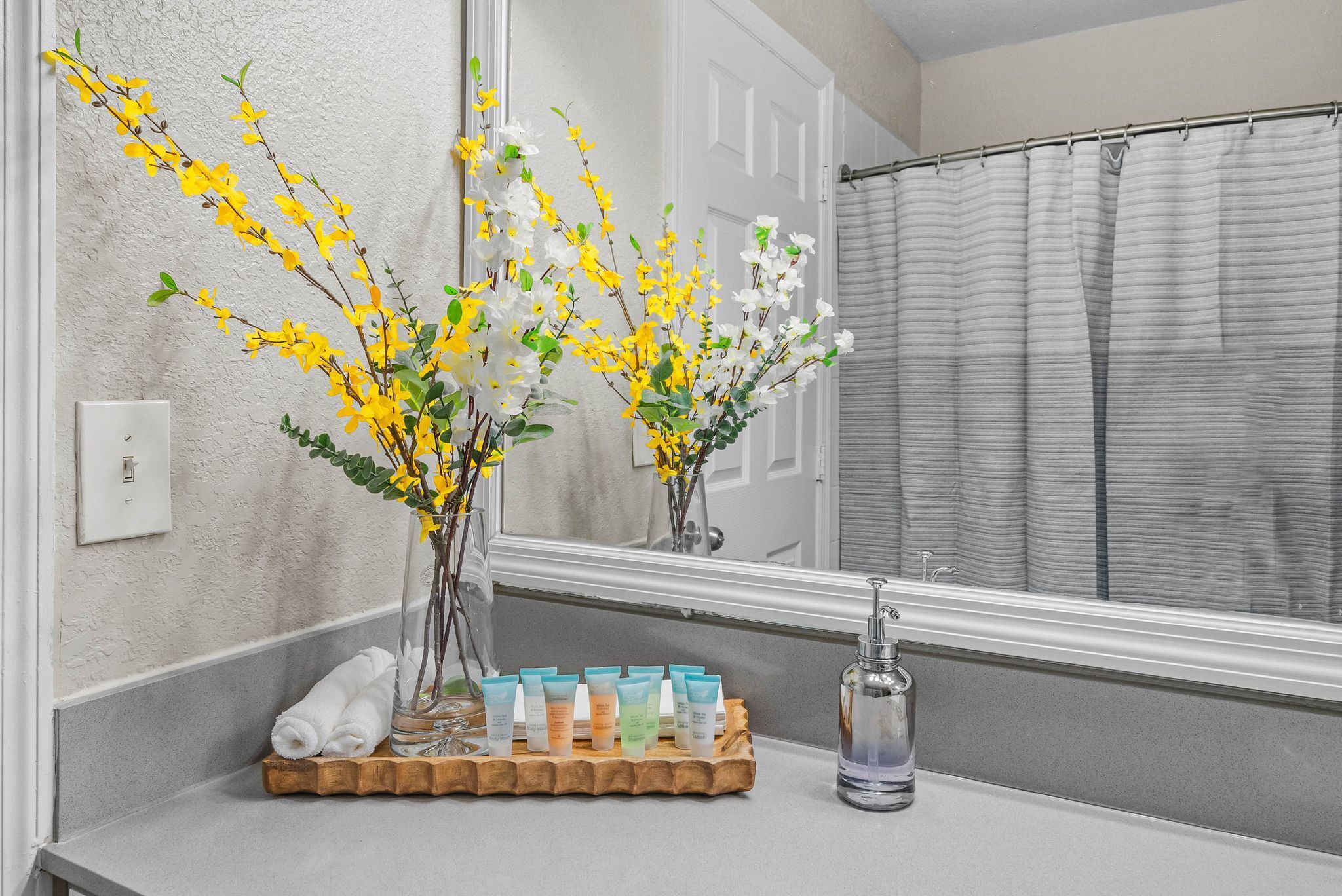 Un baño con espejo, cortina de ducha, un florero con flores y artículos de aseo en una bandeja.