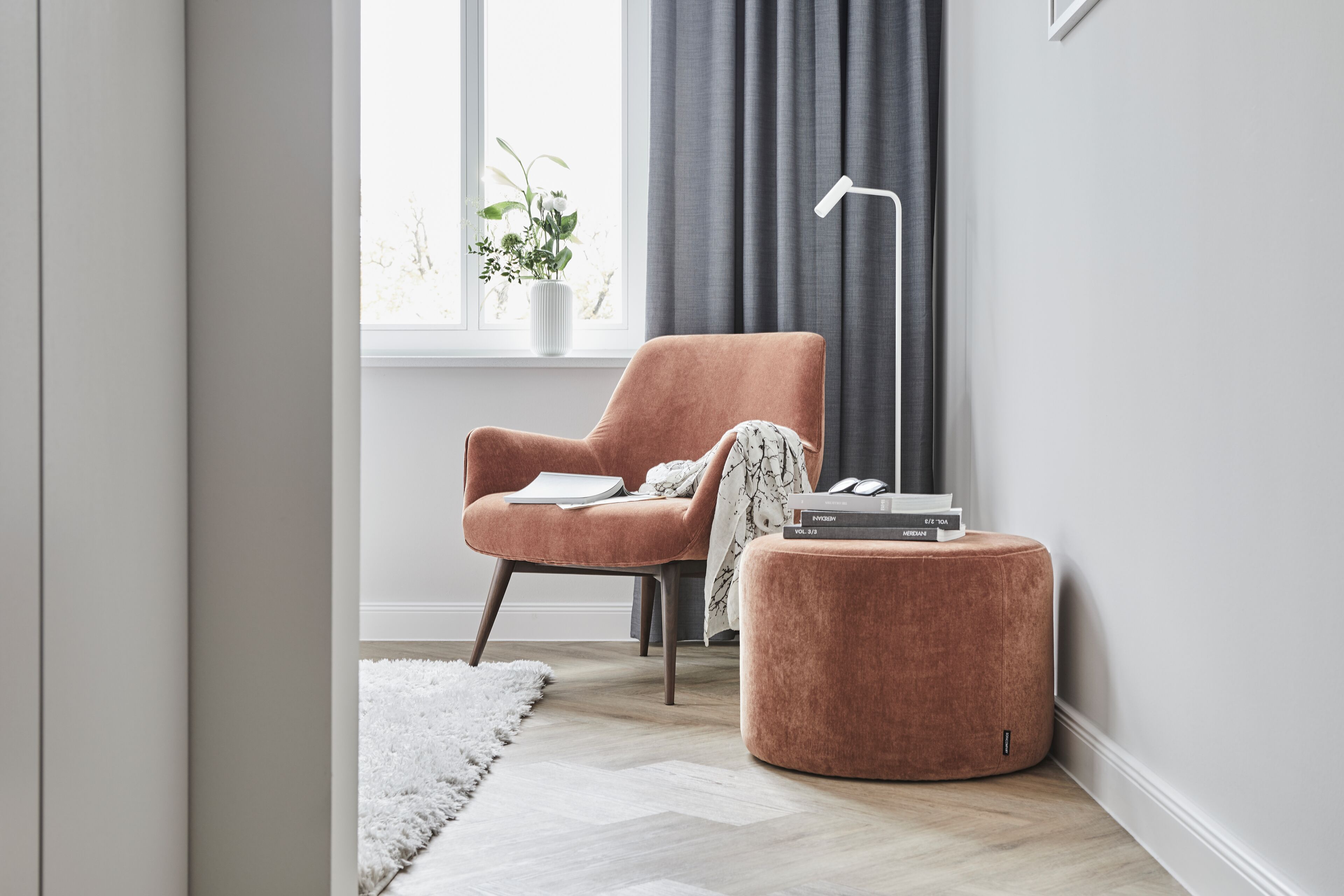 A modern living room with a plush orange armchair, a small ottoman, a white rug, and a side table with a book and a lamp.