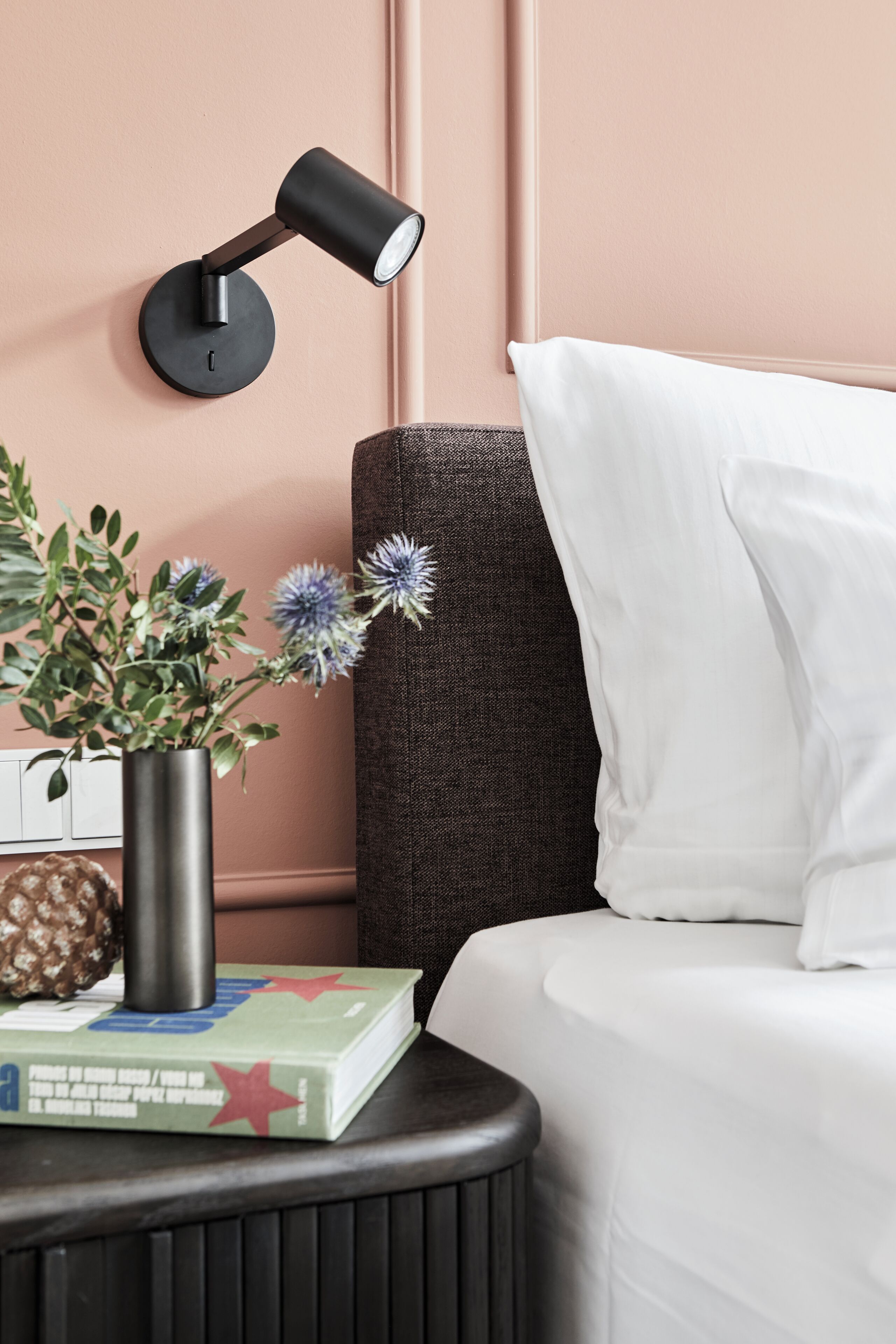 A neatly arranged bedroom with a bed, a bedside table with a lamp, a vase with flowers, and a book.