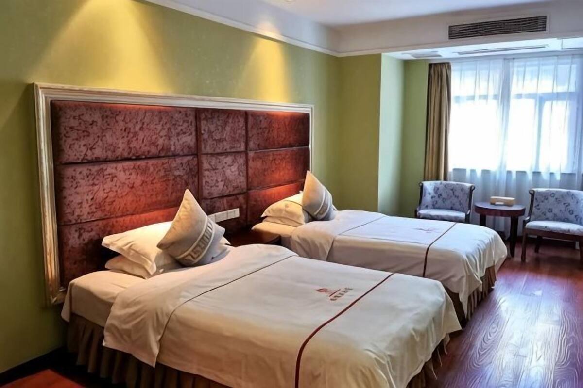 Quarto de hotel com duas camas, uma cabeceira grande, uma cadeira e uma mesinha.