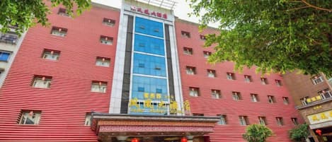 A multi-story hotel with a red facade, a central blue glass elevator, and a row of parked cars in front.