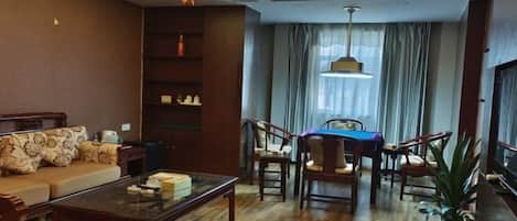 Sala de estar con un sofá, una mesa de centro y una mesa de comedor con sillas. Hay una estantería y una planta al lado derecho.