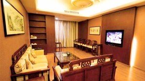 Habitación de hotel con sofá, banco de madera, televisor, mesa y estantería.