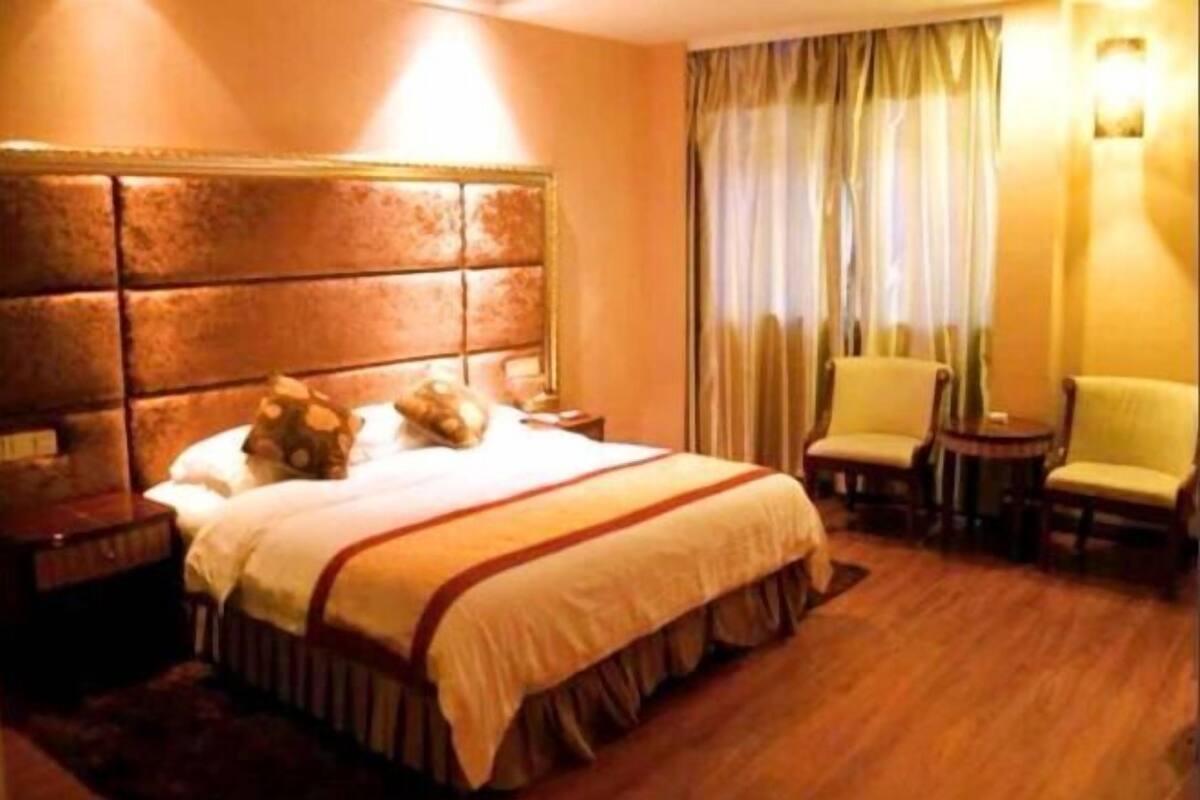Quarto de hotel com uma cama grande, cabeceira, duas cadeiras, mesa de cabeceira e janela com cortinas.